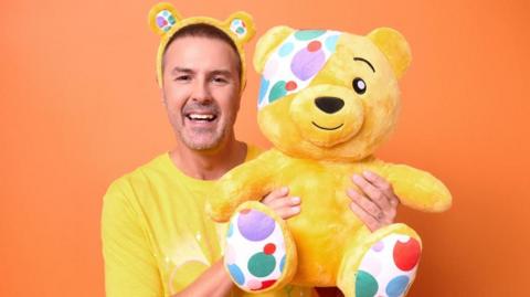 A man wearing a yellow tshirt and bear ears, holding a yellow bear with a spotted eye patch. He is smiling standing in front of an orange background.