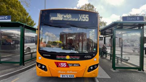 The front of a yellow bus. In the sign at the top it says 'Norwich express via Coltishall X55'. Either side of the bus are bus shelters.