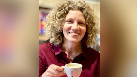 Lynda Shahwan is smiling. She has long curly blonde hair and is wearing a burgundy coloured blouse with a gold beaded necklace. She is holding a cup of coffee.