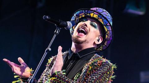 Boy George, wearing  a multi-coloured jacket and hat, claps along to music and is singing into a microphone.