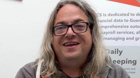 A man with long grey hair and black glasses is wearing a grey hoodie and T shirt he is in front of a TCS poster
