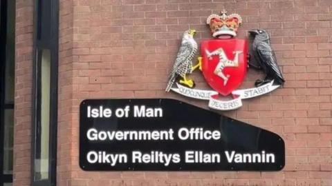 The out side of the government building, which has a black sign that reads Isle of Man Government Office with a Manx translation underneath, below the government emblem, which has the Three Legs of Man on shield between two birds.