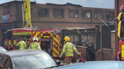 A burned-out building in the background with a fire engine and firefighters wearing safety gear in the foreground