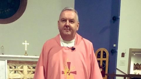A priest, who is an older man with short grey hair, is smiling and standing at the front of a Catholic church. He is wearing a pink robe which has a cross on it. 