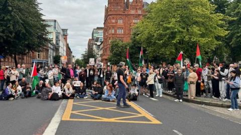 A large group of people sitting across a main road. They are holding red, green and black Palestine flags.