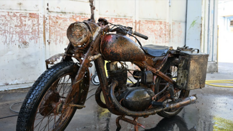 A rusted but largely well-preserved World War Two-era Wehrmacht motorcycle pictured parked in a building resembling a warehouse.