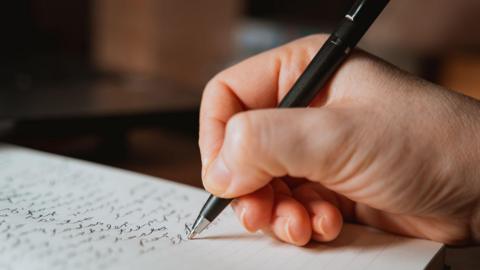 A stock image of a close up hand holding a pen and writing on a piece of paper. 