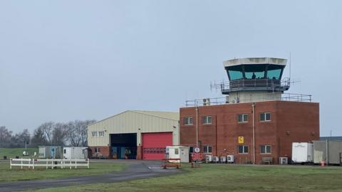 A view of the control tower at Coventry Airport showing a glass hexagonal tower on top of a red brick building, which is next to a big metal hanger.