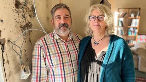 Tony and Becs Wadley are standing in the living room of their Gower Peninsula home. On the wall next to them there is black mould and an exposed electrical socket after internal wall insulation was badly installed.