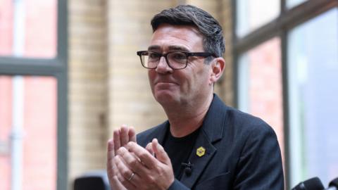 Andy Burnham gesticulates on stage at the People's History Museum in Manchester. He wears a navy jacket over a black t-shirt and black spectacles.