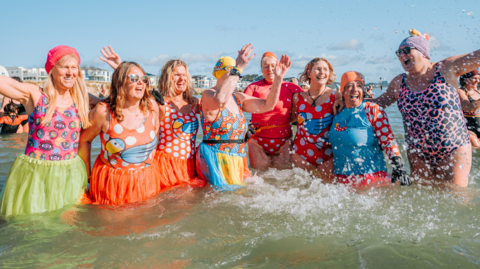 Eight women wearing colourful costumes cheer as they stand thigh-deep in the sea at Sandbanks