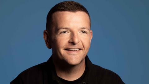 Studio-style head and shoulders shot of a smiling Kevin Bridges. he is wearing a black top and has been photographed against a blue backdrop.