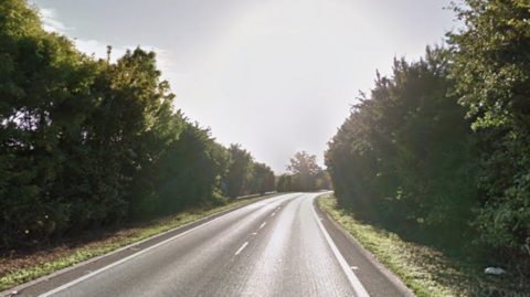 An empty stretch of A-road. There are trees on either side of the road and bright sun. 