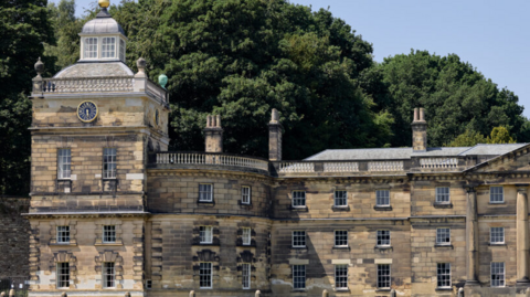 The south end of the vast Wentworth Woodhouse frontage, the brick work is light sand stone in colour and set over three floors. Each floor has 9 windows. Towards the left side of the image there is a rounded turret like section of the building. At the southern tip stands the tower, complete with a white glass roof feature and a clock just under the roof space facing outwards. Three chimneys are visible and in the background is a blue sky above large mature trees.