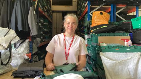 A woman wearing a white T-shirt and red lanyard stands at a table in a small room with shelving behind her. On the shelves are school blazers, trousers and green crates of uniform stacked.