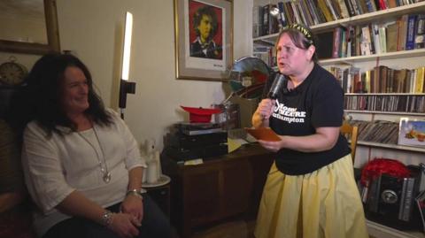 An actress, lit by a portable light stand, performs in a darkened living room. She is speaking into a pretend microphone, as if pretending to be a news reporter. A woman in the audience is laughing. Behind the actress are bookshelves and other home furniture.