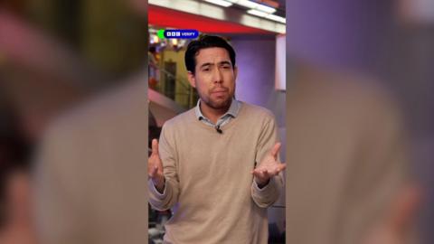 A male journalist wearing a light brown jumper talks to the camera