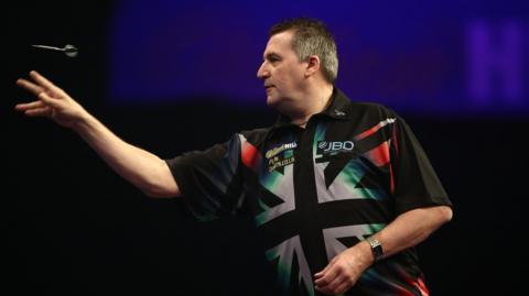 Ronnie Baxter seen side on throwing a dart with his right hand. He is wearing a black shirt with a black and white Union Flag on it.