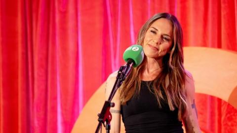 A woman with wavy brown hair and tattoo on her arms stands in front of a microphone, against a red curtained backdrop. 