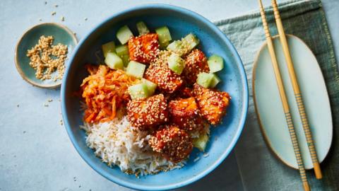 A blue bowl filled with cubes of salmon and cucumber with shredded carrot and rice on a blue surface with a pair of chopsicks