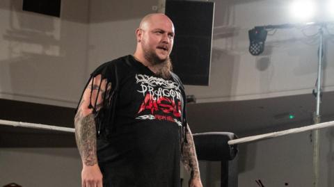 Dragon Aisu, real name Dante Richardson, wearing a black top with his wrestling name on it. He has tattoos on his arms. He has a beard. He is standing in a wrestling ring.
