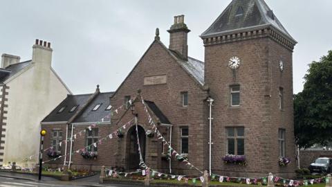 The image shows St John's Parish Hall pictured on a cloudy morning. There are are strings of bunting from the entrance.
