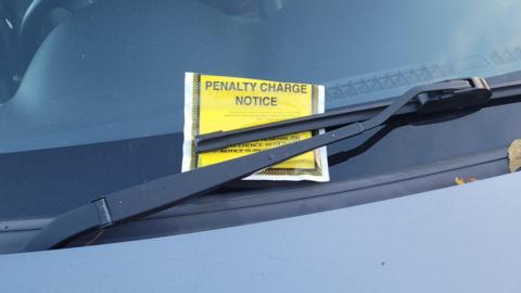 A close-up of a yellow penalty charge notice placed behind the windscreen wiper of a car.