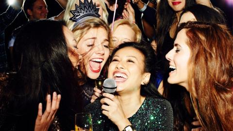 A group of women singing karaoke together, smiling and holding drinks