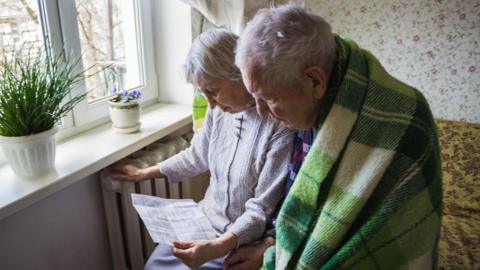 Two pensioners sat near a radiator looking at heating bills. One of them has a blanket around him. 