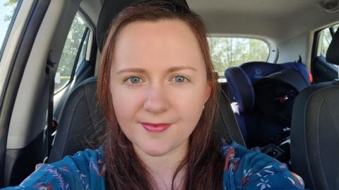 Rachel has taken a selfie photo in her car. She has shoulder length brunette hair and is wearing a blue blouse top. 