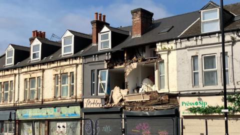 A row of shopfronts, with a building in the middle collapsed, with the front exposing rooms inside.