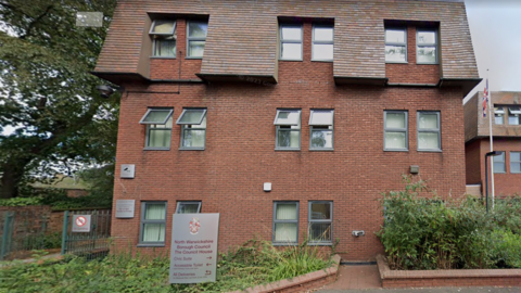 A red brick building with a roof and dozen of windows. Bushes and brick walls line the front of the property. A Union Jack flag can be seen towards the right hand side of the building.