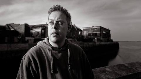 Nick Quantrill. A moody black and white photograph taken outside with a boat in the background.  He wears a hoodie and glasses.