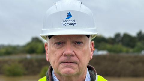 Martin Fellows looks into the camera. He wears a white hard hat with the words National Highways written on it. He also wears a florescent green coat. Behind him trees can be seen.