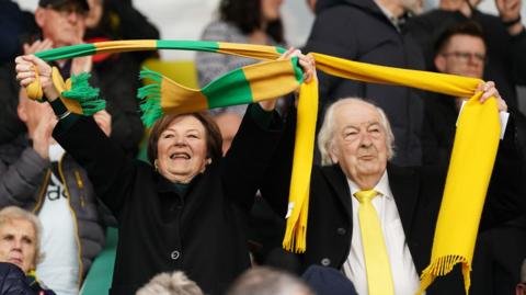 Delia Smith and Michael Wynn-Jones holding yellow and green scarves in the air while in the stands of a football stadium