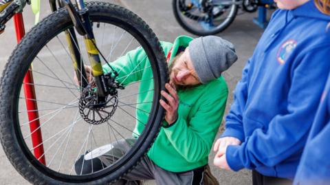 A man wearing a green hoodie and grey beanie hat is looking at a bike tyre. 