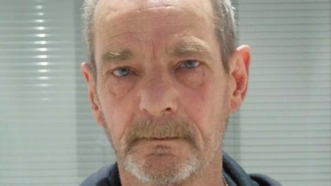 A mugshot of a man with a grey goatee, grey hair and short beard. He has blue eyes and looks into the lens with a neutral expression.