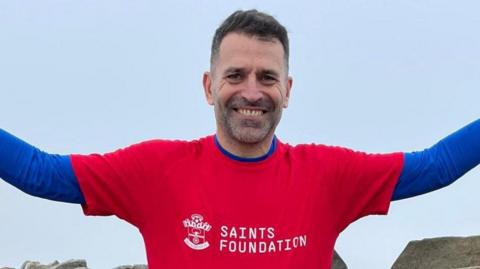 Franny Benali smiling with his arms out as he looks at the camera wearing a Saints foundation t-shirt.