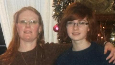Woman on the left is wearing glasses and has long ginger hair. She is also wearing a brown jumper with a long gold necklace around her neck. She has her arm around a younger woman who is wearing glasses and short ginger hair in a bob and is wearing a blue jumper.