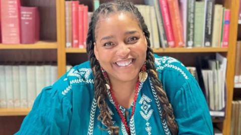 Désirée Reynolds has long, brown braided hair and is wearing a turquoise top with white stitching across her shoulders and down her chest. She is smiling at the camera as she sits in front of a bookshelf.