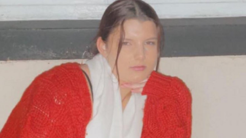 Tamzin Hall is seen as a young teenager. She is wearing a red cardigan and a white scarf. She is holding her hand under her chin and looking at the camera.