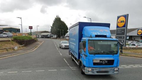 A blue lorry prepares to turn into Callington Road at the busy junction with Gilston Road on the edge of a retail estate.