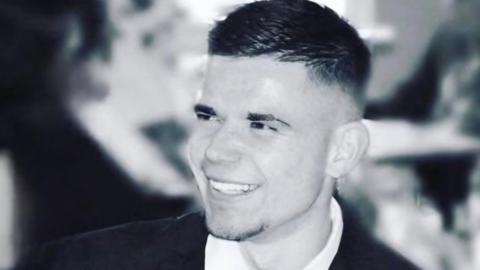 A black and white photo of Mason Jones, a 30-year-old man, wearing a suit and tie and smiling 