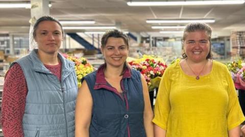 Four people stand in a row in a flower packing warehouse. Three women from left to right wearing a blue vest, a navy vest, bright yellow top, and a man in a navy polo at the end. The flowers behind them are wrapped in yellow paper, and coloured pink, white and red.