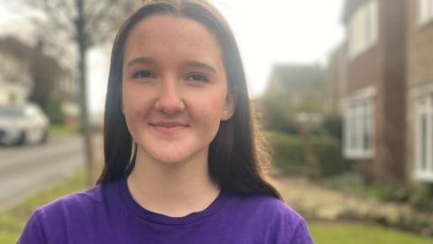 A teenage girl with brown hair, wearing a purple t-shirt. She is standing outside a property.