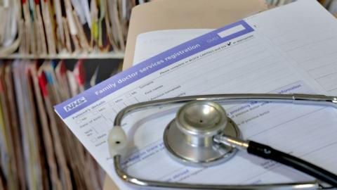 A stock image of an NHS document with a stethoscope lying on it. In the background are files on a shelf.