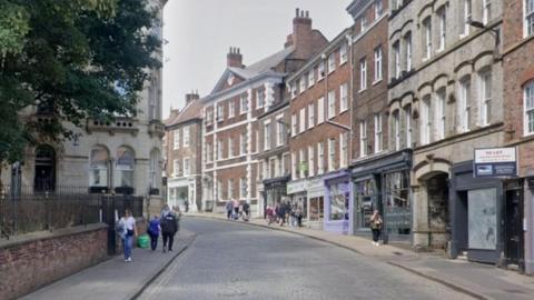 The image shows a gently curving, cobblestone street lined with historic brick and stone buildings. Many of the buildings have large ground‑floor shop fronts. People are walking along the pavement on both sides of the street. On the left side, there is a stone building partly hidden behind railings and trees.