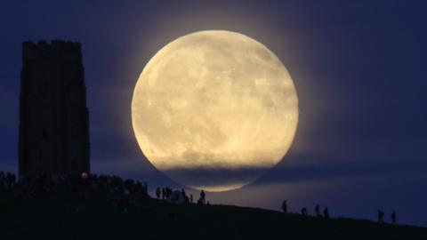 A Strawberry Moon rising above Glastonbury Tor in Somerset. The moon is large and bright, despite some cloud cover at the bottom of it. The silhouette of Glastonbury Tor can be seen to the left of the frame and there are people at the bottom of the frame. They are standing on the hill.
