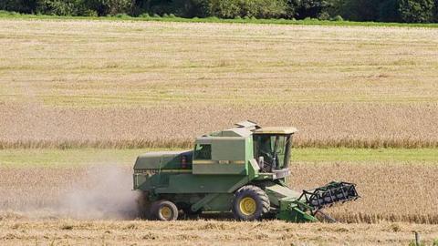 A combine harvester on a field. There are trees in the background and lots of green space. 