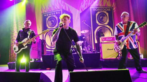 Frank Carter (centre) performs with Glen Matlock and Steve Jones of the Sex Pistols on stage at O2 Forum Kentish Town. The backdrop is a graphic of two huge speakers, branded "nowhere" and "boredom".